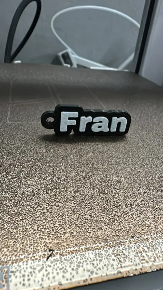 Llavero impreso en 3D con el nombre Fran en letras blancas sobre base negra, apoyado sobre la cama de la impresora