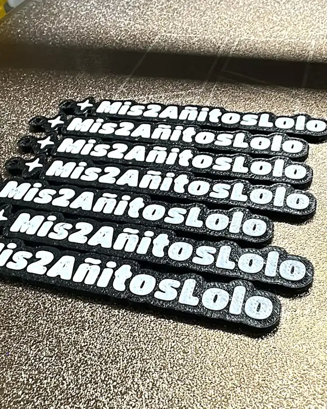 Siete llaveros con el texto Mis2AñitosLolo y una estrella, recién impresos sobre la cama de la impresora 3D