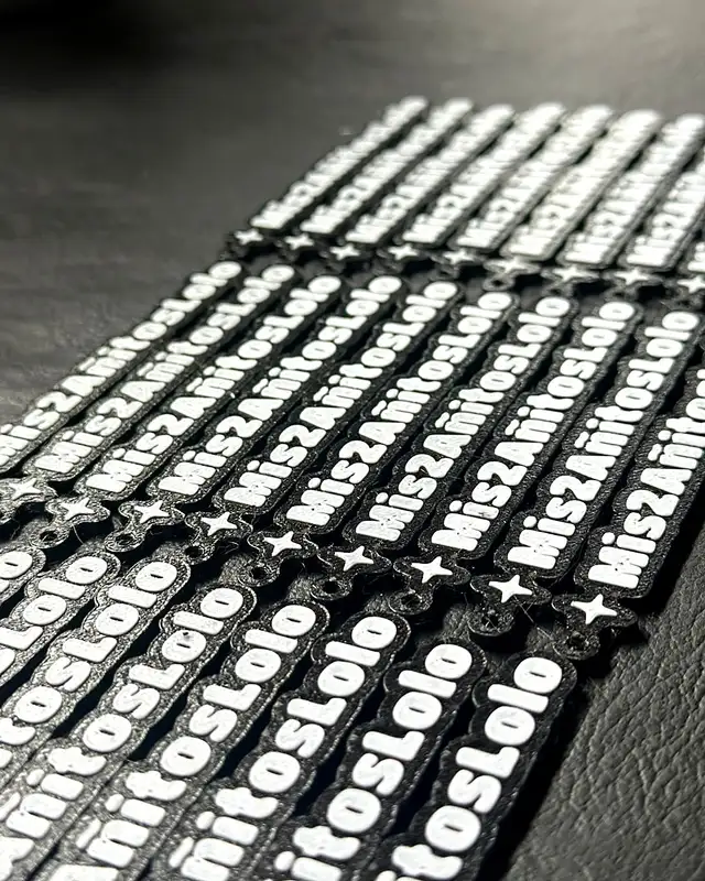 Decenas de llaveros impresos en 3D con el texto Mis2AñitosLolo en blanco sobre base negra, con una estrella, alineados sobre una mesa